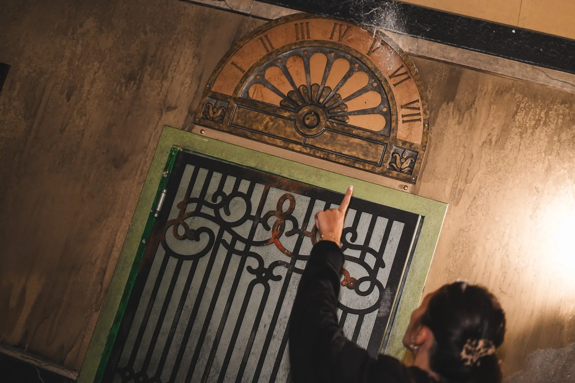 Joueuse qui désigne une grille ornée sous une horloge ancienne, dans une salle d'escape game Game Over